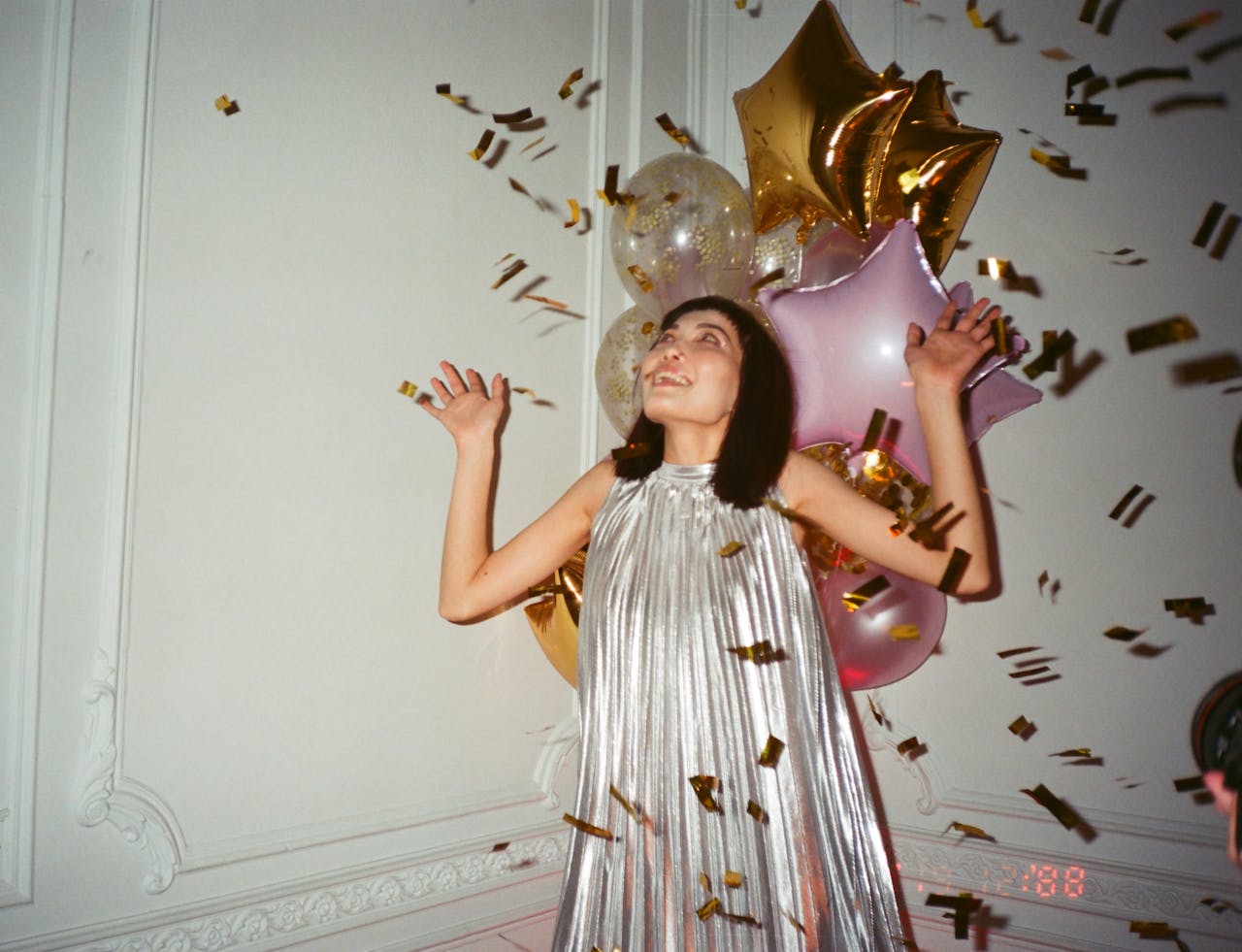 A woman in a silver dress celebrates with balloons and confetti indoors.